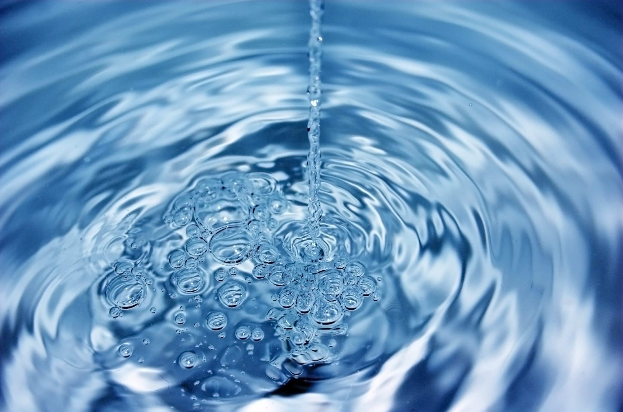 A tall glass is being filled with water, creating bubbles and splashes, with ice cubes melting on a wet wooden surface and a blurry green background.